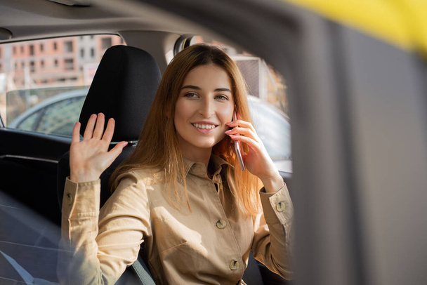 Young businesswoman behind the driver's seat talking on the phone while stopping and waving hello through the window - Фото, изображение