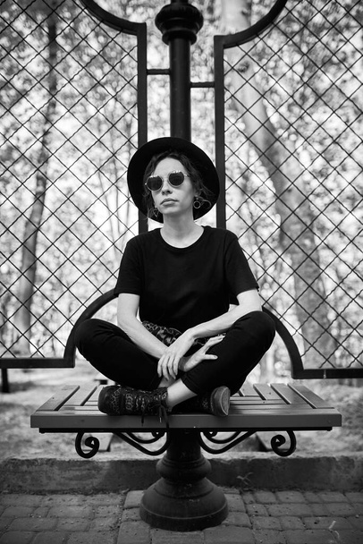 The girl in black clothes, a hat and sunglasses is sitting on a park bench - Foto, Bild