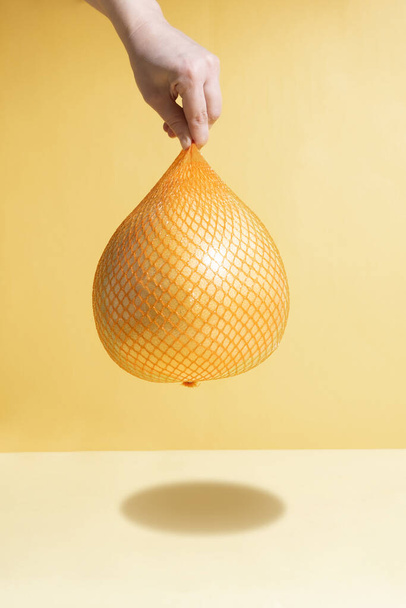 Female hand hangs yellow fresh pomelo in mesh packaging on a yellow and orange background in the sunlight, close up. Modern isometric concept. - Foto, Imagem