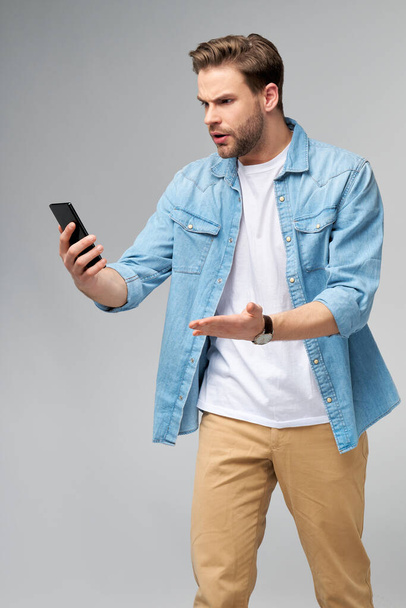 Young shocked stressed angry and frustrated man using smartphone over grey background - Φωτογραφία, εικόνα