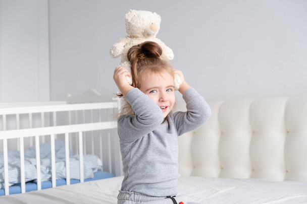 little girl in grey turtleneck playing with teddy bear on white bedding. - Photo, Image