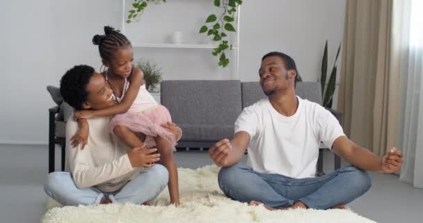 Afro amerykańska młoda kobieta mama i czarny mężczyzna tata siedzieć boso na podłodze w salonie w przytulnym domu medytować mało słodkie etniczne córka aktywna dziewczyna kolidować z rodzicami robi jogi ćwiczenia uściski matka - Materiał filmowy, wideo