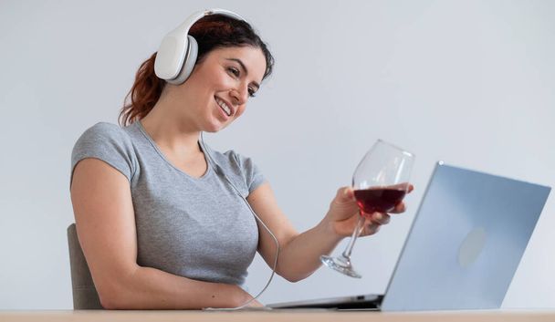 Een vrouw in een koptelefoon drinkt rode wijn en communiceert met vrienden via videocommunicatie op een laptop. Online chatten in quarantaine - Foto, afbeelding