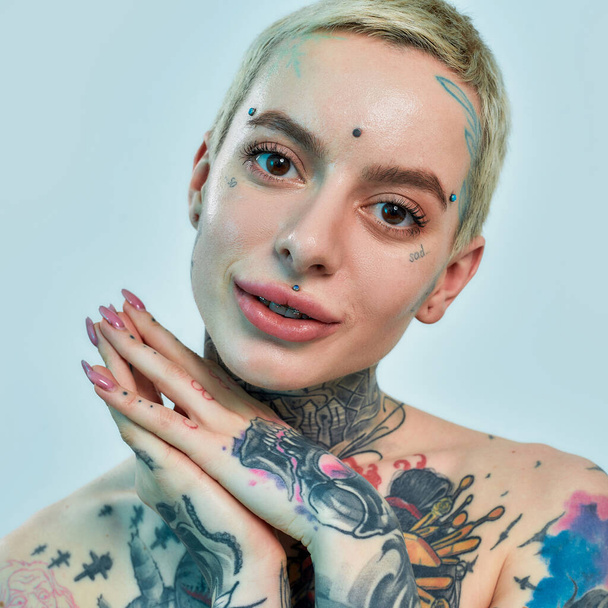 Tattoo and piercing. A portrait of a white tattooed woman with piercing looking into a camera with her hands together near her head slightly tilted - Photo, Image