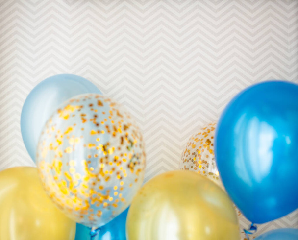Festive decoration of a bright room with blue and transparent balloons with golden confetti. We celebrate the holidays at home. Bouquet of balloons on the background of wallpaper with zigzags - Fotografie, Obrázek