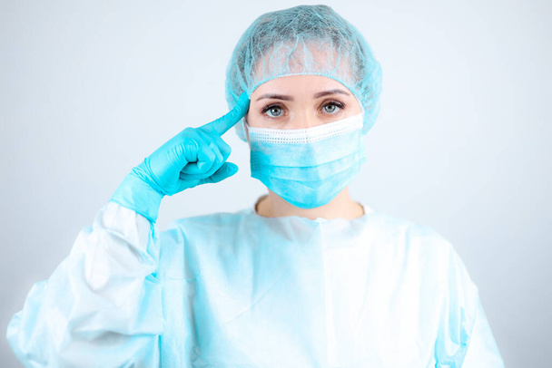 μια νοσοκόμα με ιατρική ρόμπα, μάσκα και προστατευτικά γάντια δείχνει ένα δάχτυλο στο κεφάλι της - Φωτογραφία, εικόνα
