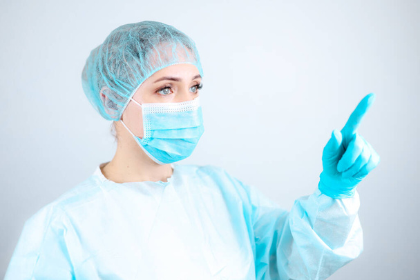 медсестра в медицинском халате, маске и защитных перчатках стоит боком и показывает пальцем вперед - Фото, изображение