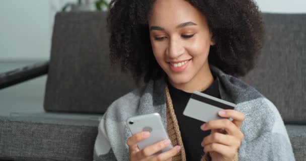 Portrait smiling afro american woman makes online purchase through mobile phone holds smartphone in her hands and credit bank card buys goods books travel ticket orders food delivery home by courier - Footage, Video