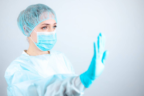 een verpleegster in een medisch gewaad, masker, en beschermende handschoenen staat zijwaarts met haar hand uitgestrekt in een "stop" gebaar. - Foto, afbeelding