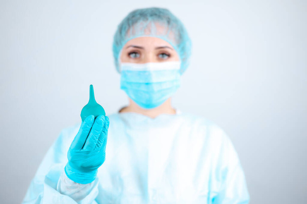 Μια νοσοκόμα με ιατρική ρόμπα, μάσκα και προστατευτικά γάντια κρατά ένα κλύσμα στα χέρια της - Φωτογραφία, εικόνα