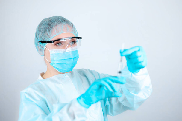 Een verpleegkundige in een medisch gewaad, masker en beschermende handschoenen met een transparante bril op haar gezicht verwijdert de dop van de naald van de spuit - Foto, afbeelding