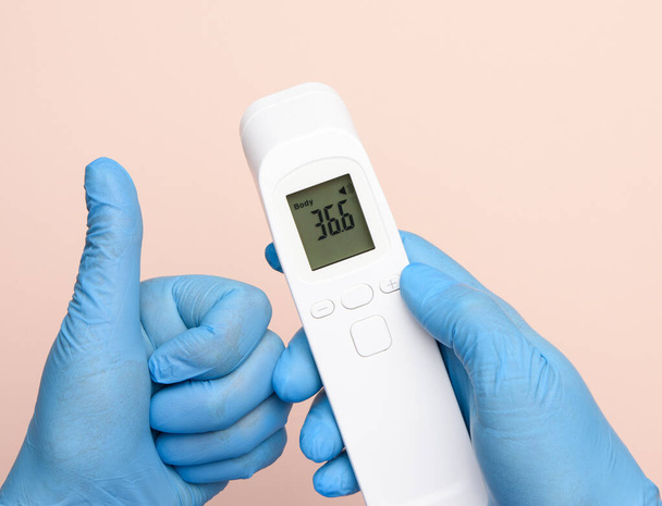manos en guantes de látex azul sostienen un termómetro electrónico para medir la temperatura, dispositivo sin contacto  - Foto, imagen