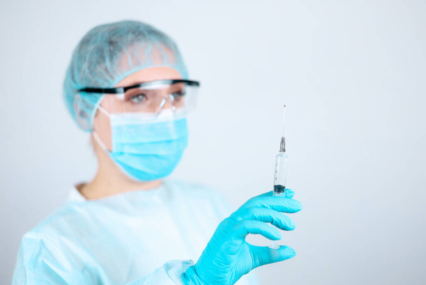 A nurse in a medical gown, mask and protective gloves with transparent glasses on her face looks at the needle of a syringe for injection - Photo, image