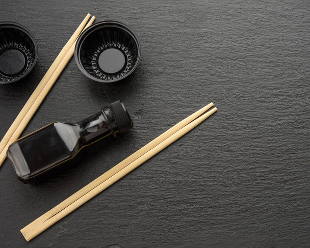 wooden sticks for sushi, bottle of soy sauce and disposable plastic plates on black background, utensils for delivery - Fotó, kép
