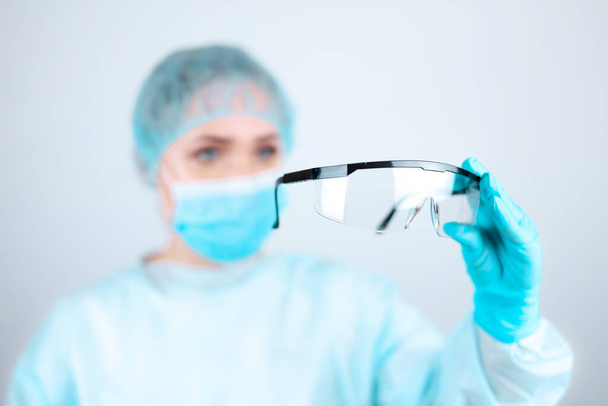 Een verpleegkundige in een medisch gewaad, masker en beschermende handschoenen kijkt in transparante glazen - Foto, afbeelding