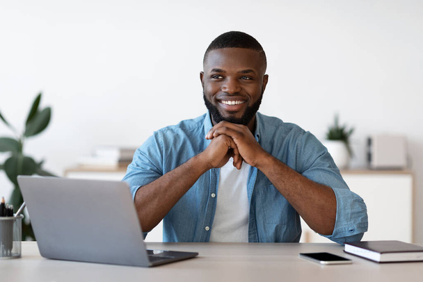 Pensive czarny człowiek biuro pracownik siedzi na biurku z laptopem i uśmiechem - Zdjęcie, obraz