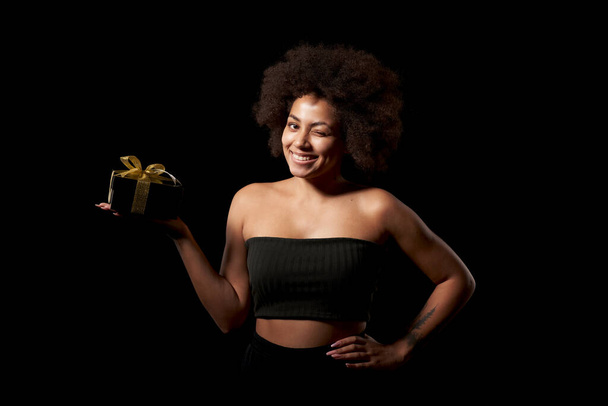 Black Friday sale, holidays gift, present, celebration concept. Happy playful afro-american female holding one black gift box with golden bow over black background, looking at camera winking - Foto, Bild