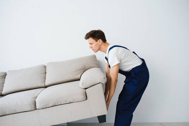 Loader moves sofa, couch. worker in overalls lifts up sofa, white background. Delivery service concept. Courier delivers furniture in case of move out, relocation. - Photo, image