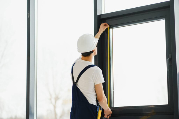 Workman in overalls installing or adjusting plastic windows in the living room at home - Photo, Image
