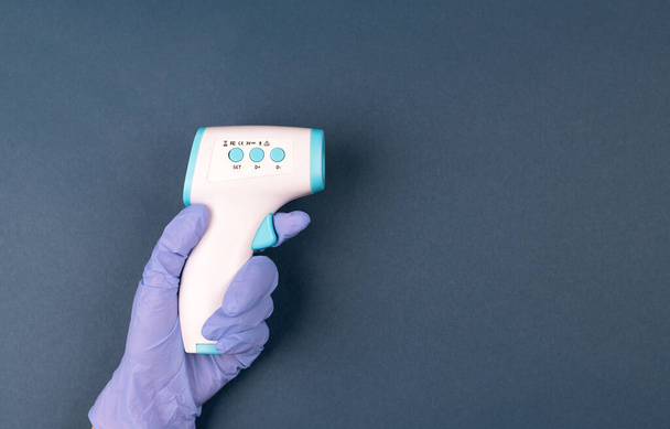 濃い青の背景に空気温度計を保持手袋で手。アンチウイルスは概念を保護します。医務室測定製品。医療ホームアクション。遠くの温度チェック. - 写真・画像