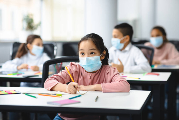 Aasialainen tyttö maskissa istuu pöydällä luokkahuoneessa - Valokuva, kuva