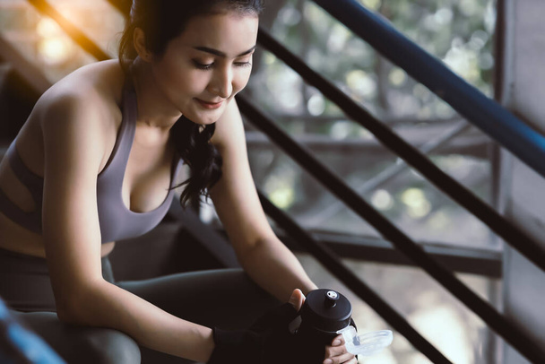 Happy sport asian woman taking a rest during exercising in gym by sitting on steps and holding bottle of water Beautiful young girl wear sport wear She get tired, training for diet Sport concept - Photo, Image