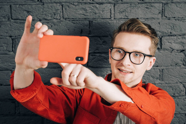 Giovane uomo gioioso in camicia rossa e occhiali scattare foto selfie sul cellulare su uno sfondo di muro di mattoni neri. Copia, spazio vuoto per il testo . - Foto, immagini