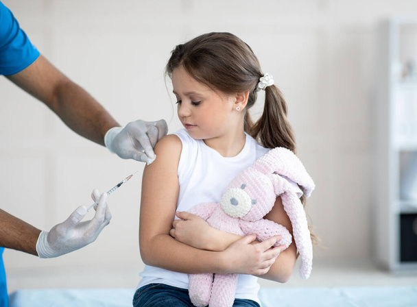 Doctor doing injection of coronavirus vaccine to little girl with toy bunny at clinic. Medicine and vaccination - Photo, Image