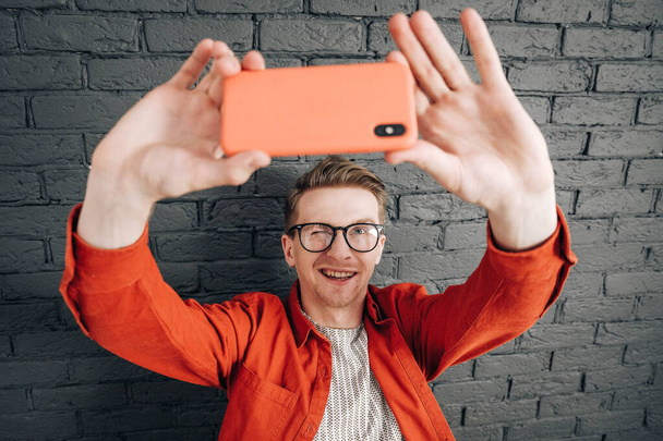 黒レンガの壁を背景に、携帯電話で自撮り写真を撮る赤いシャツと眼鏡の青年。テキストのコピー、空のスペース . - 写真・画像
