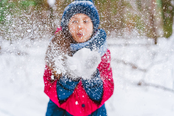  Mała dziewczynka wieje śnieg rękawiczkami na tle płatków śniegu bokeh. Dziecko bawi się na świeżym powietrzu w zimowym lesie - Zdjęcie, obraz
