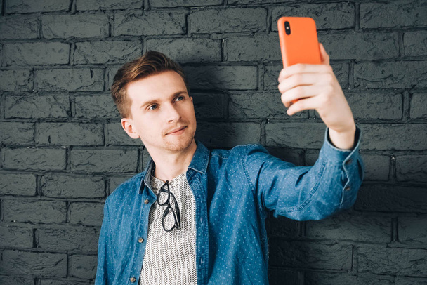 Young joyful man in blue shirt and glasses taking selfie photo on cellphone on a background of black brick wall. Copy, empty space for text. - Photo, Image