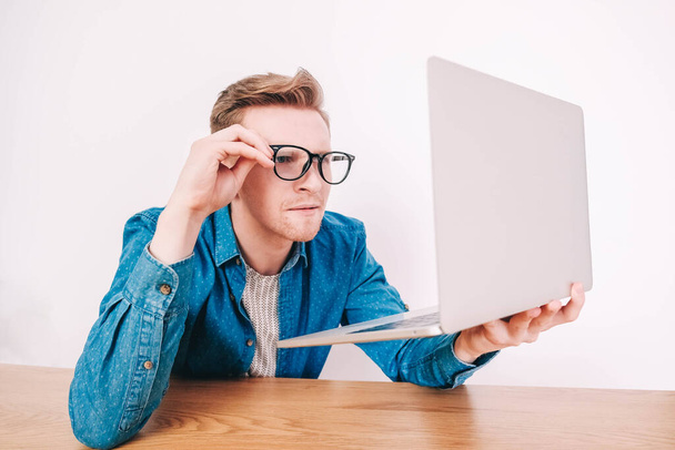 Young man in shirt and glasses he looks at the laptop in amazement and with a funny face sitting at the table on a white background. Funny face and amazement. Copy, empty space for text. - Foto, Bild