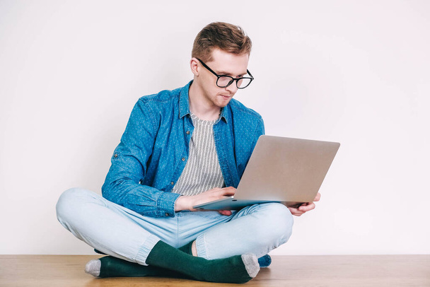 Mladý muž v košili a brýlích pracuje na notebooku, který sedí na stole na bílém pozadí. Práce na volné noze. Kopírovat, prázdné místo pro text. - Fotografie, Obrázek