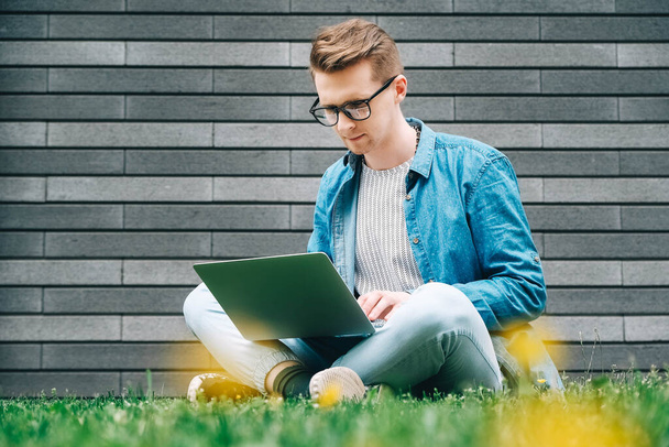 Young man with glasses businessman hipster sitting on green grass and using laptop computer on a gray wall background. Freelance work concept. Copy, empty space for text. - Foto, immagini