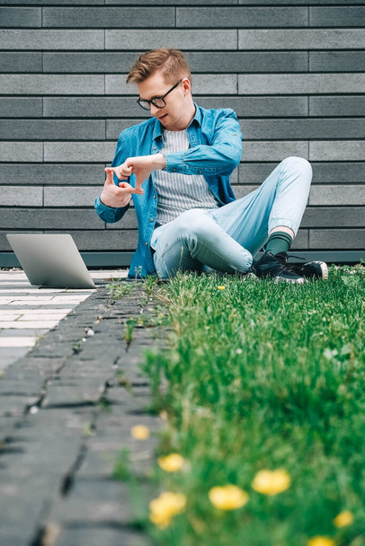 緑の芝生の上に座って、灰色の壁の背景にラップトップコンピュータを使用してメガネビジネスマンのヒップスターを持つ若い男。フリーランスの仕事のコンセプト。テキストのコピー、空のスペース. - 写真・画像