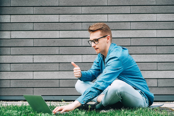 Young man businessman in shirt, jeans and glasses sitting on green grass and using laptop computer on the background of a gray wall of the house. Freelance work concept. Copy, empty space for text. - Photo, Image