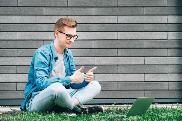 Jonge man zakenman in shirt, jeans en bril zittend op groen gras en met behulp van laptop computer op de achtergrond van een grijze muur van het huis. Freelance werk concept. Kopieer, lege ruimte voor tekst. - Foto, afbeelding