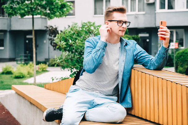 Jeune homme beau en chemise, jeans et lunettes en utilisant communique sur un smartphone assis sur un banc sur un fond de maisons. Copier, espace vide pour le texte. - Photo, image