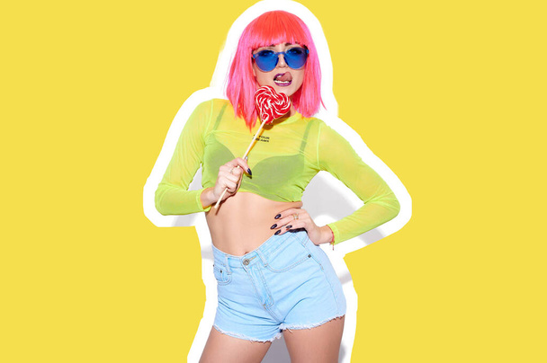 Freaky girl in bright pink wig and denim shorts lick red lollipop heart shape and show tongue on yellow background - Fotoğraf, Görsel