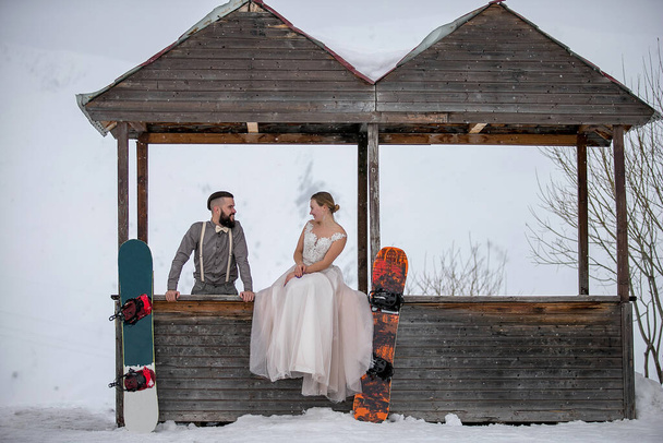 Nevěsta sedí na dřevěném domě ve svatebních šatech v blízkosti ženicha vedle jejích snowboardů a dívá se na sebe - Fotografie, Obrázek