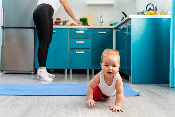 Smiling caucasian baby crawls on the floor. In the background is a sports mat and a woman stands at the kitchen shelves. - Foto, Imagem