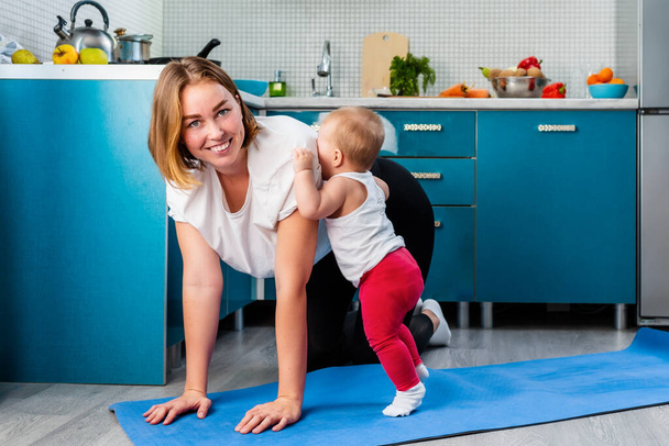 Home yoga. A young Caucasian mother do yoga sports in her kitchen while her toddler walks around. The concept of sports activity with kids. - Foto, Imagem