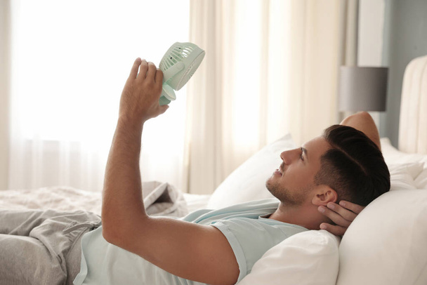 Man enjoying air flow from portable fan on bed in room. Summer heat - Foto, afbeelding