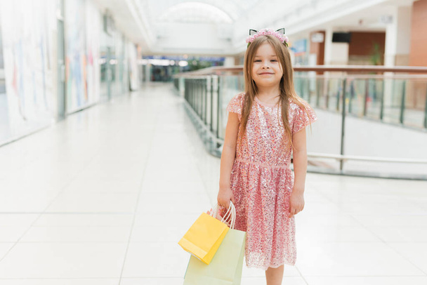 Büyük bir alışveriş merkezinde paketleri olan küçük bir moda kızı. Dükkanda poz veren güzel gülümseyen küçük kız. Alışveriş merkezinin yanındaki pembe elbiseli çocuk eğleniyor. - Fotoğraf, Görsel