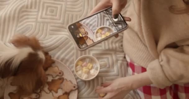 Een jong meisje met haar hond zit op een bed met een schotel peperkoek en een kopje cacao met marshmallows en fotografeert eten op een mobiele telefoon. Uitzicht van bovenaf - Video