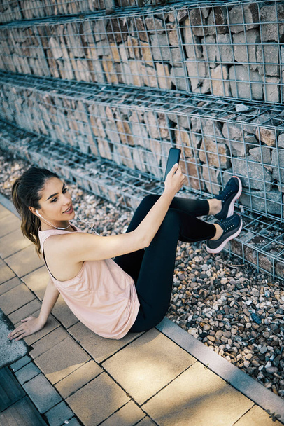 A female runner sitting on sidewalk, taking a break and using phone. - Фото, зображення
