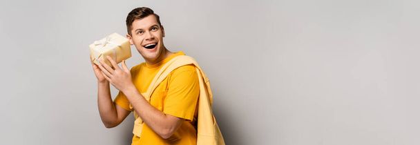 Amazed man in yellow clothes holding present on grey background, banner  - Photo, Image