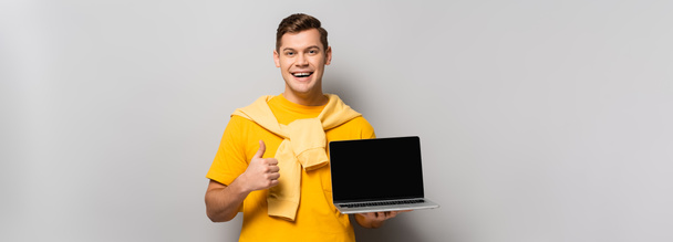 Mężczyzna trzyma laptopa z pustym ekranem i pokazuje kciuk do góry na szarym tle, baner  - Zdjęcie, obraz