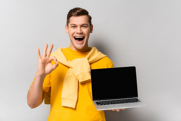 Positive man holding laptop with blank screen and showing ok gesture on grey background - Foto, imagen