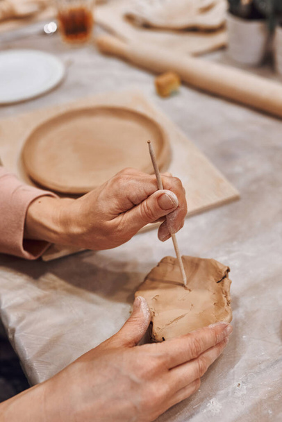Ένας κεραμίστας σε εργαστήριο κεραμικής φτιάχνει σχέδια σε πήλινο πιάτο χρησιμοποιώντας εργαλεία. - Φωτογραφία, εικόνα